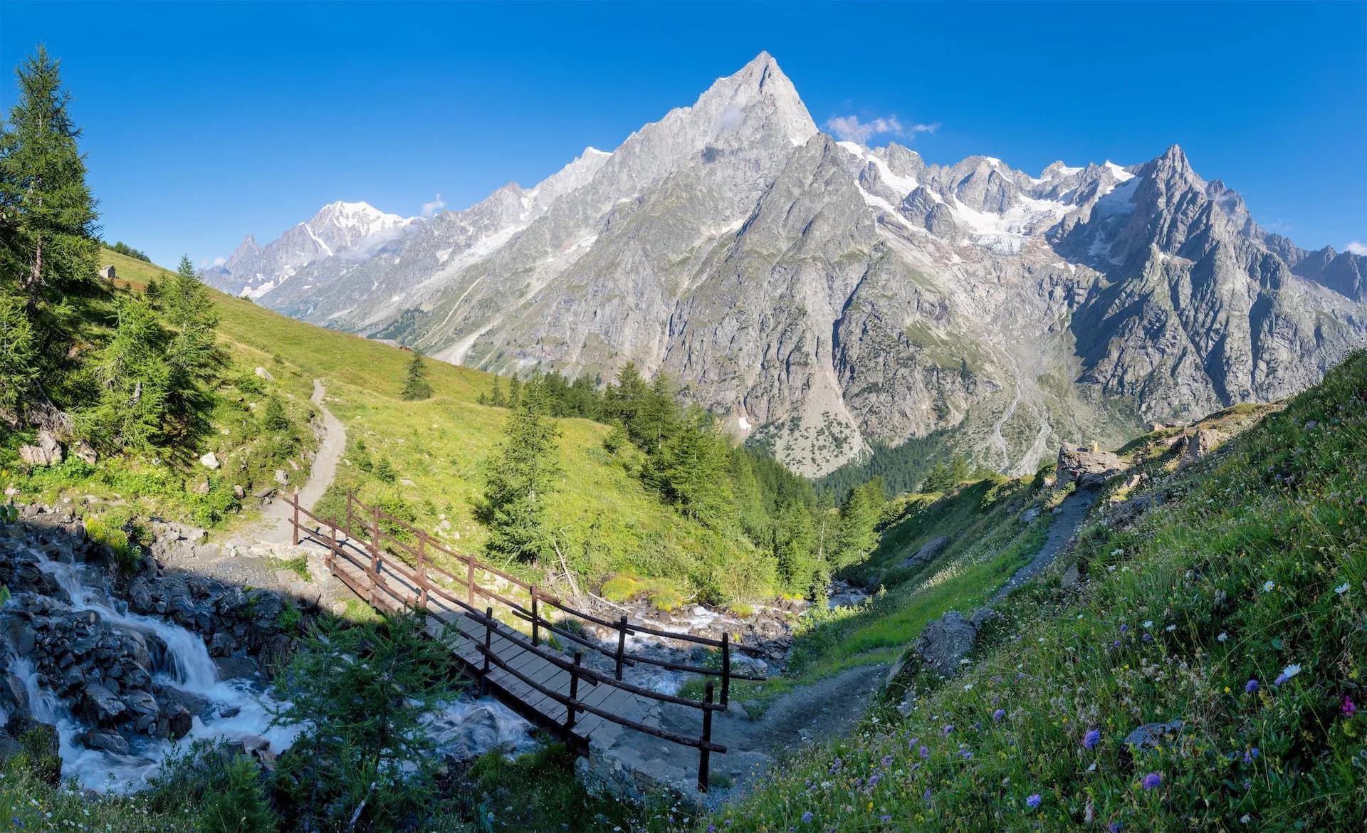 Hiking trail with wooden bridge over stream in Val Ferret valley below snow-capped Grand Jorasses massif.