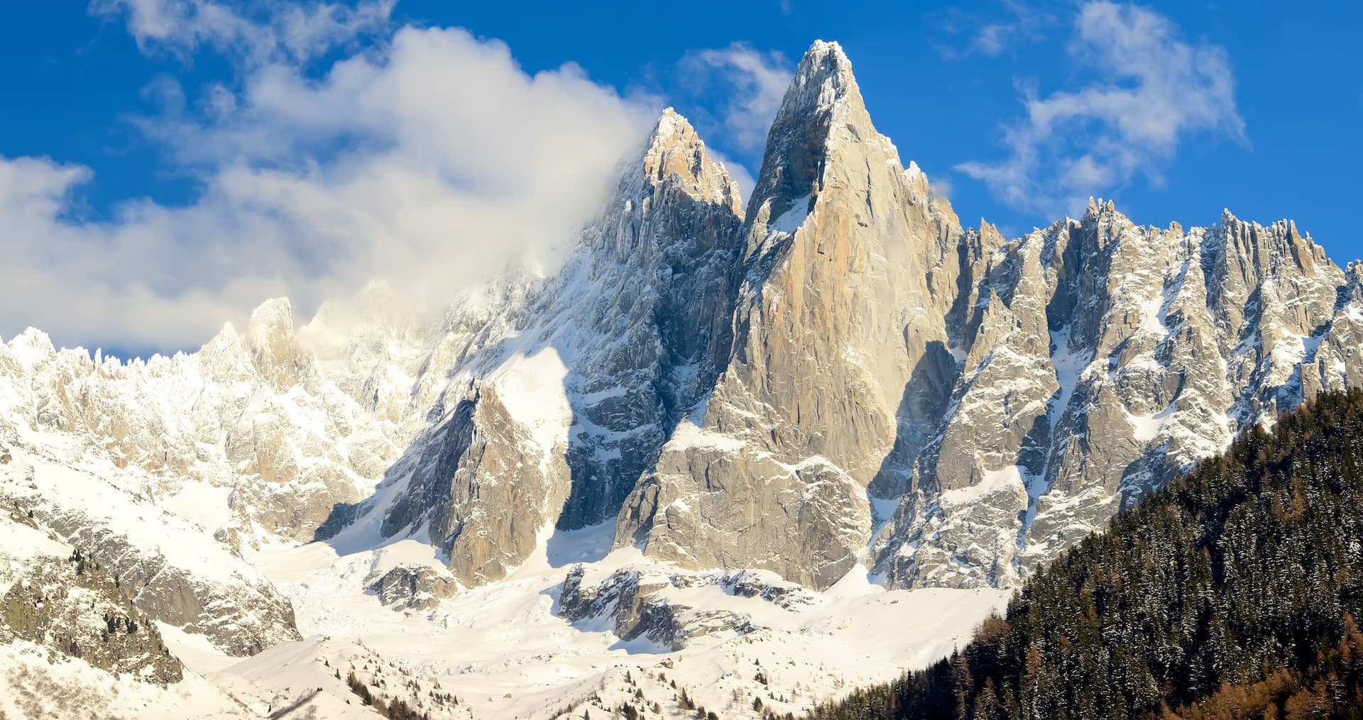 Snow-covered Aiguille du Dru mountain peaks rising above dark pine forest under blue sky with clouds.