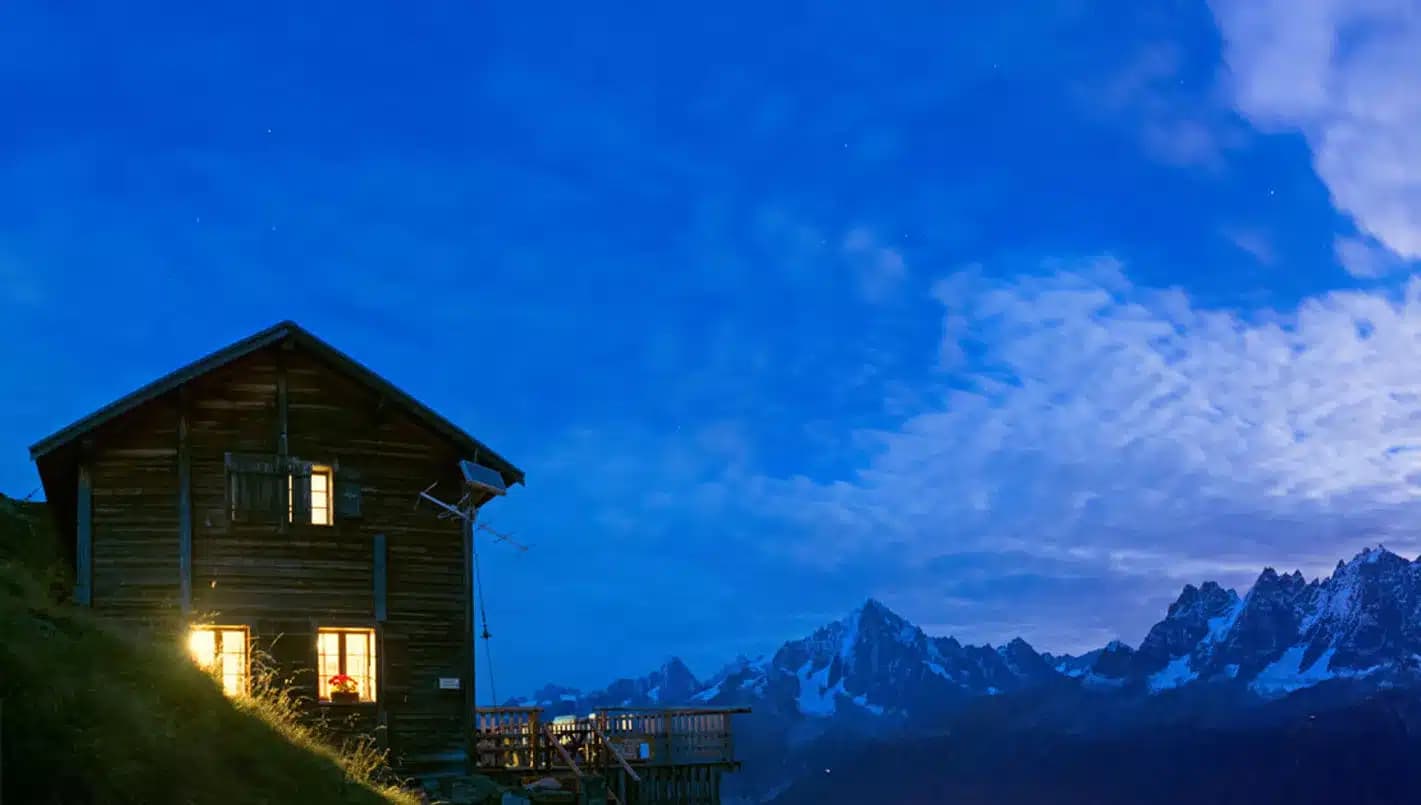 Illuminated wooden refuge on grassy slope with snowy mountains under blue twilight sky, Refuge Bellachat.