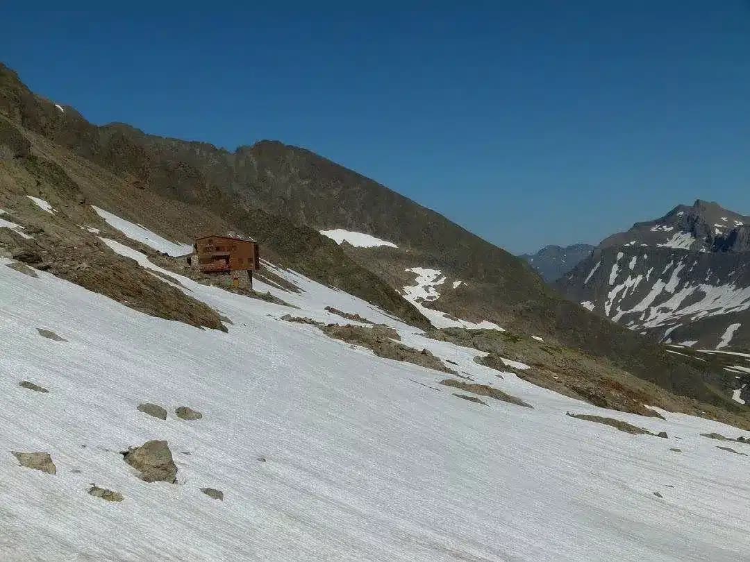 Mountain refuge hut on steep, partially snow-covered slope under clear blue sky, Robert Blanc.
