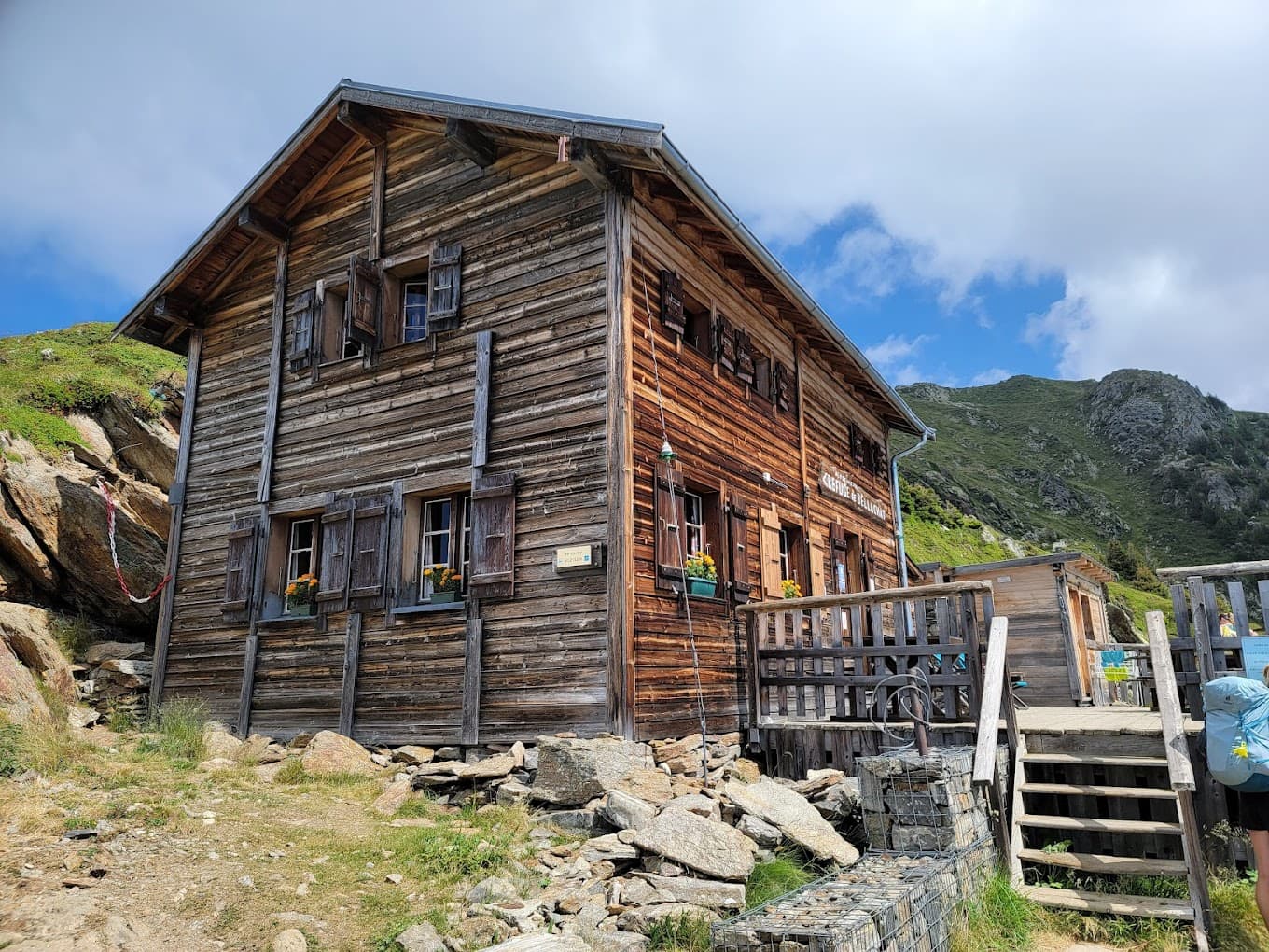 Wooden refuge building, Refuge Bellachat, nestled against green mountainside under blue sky