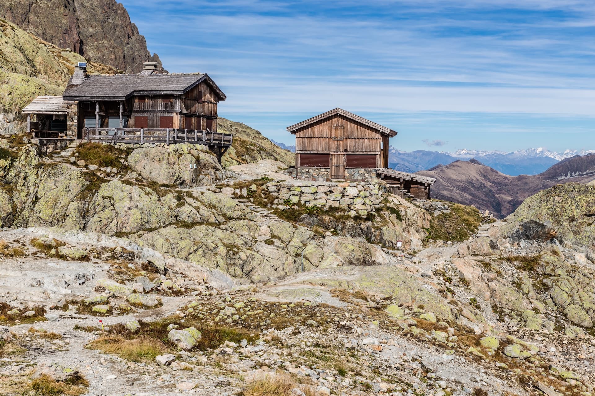 Wooden mountain huts on rocky terrain with distant snow-capped peaks under a blue sky.