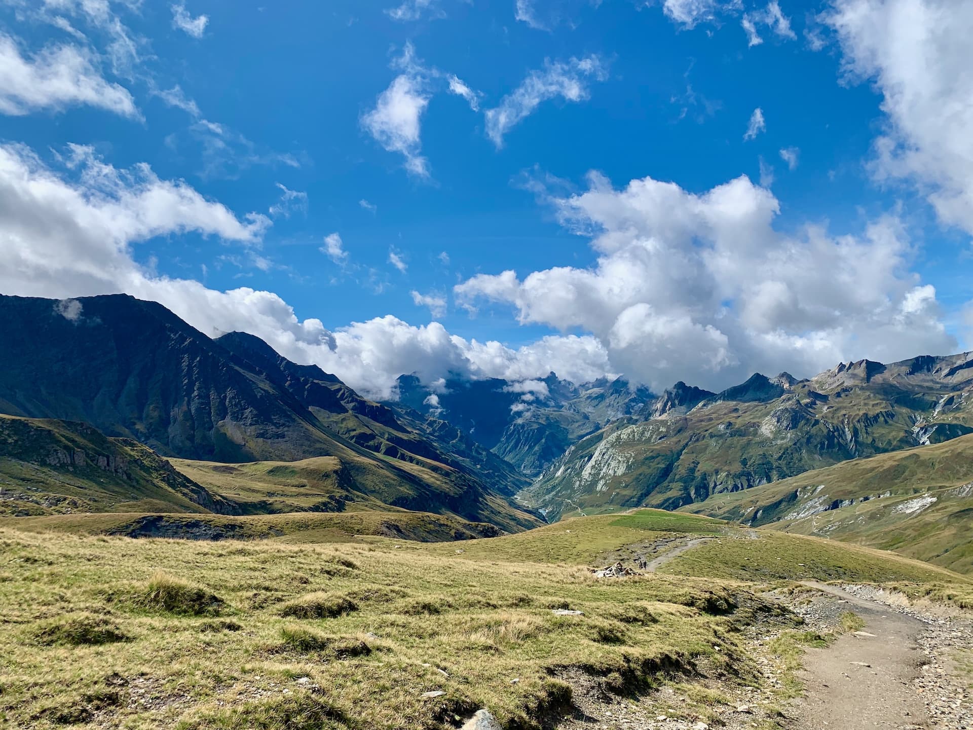 Hiking trail on grassy slope toward Col de la Seigne with dramatic mountains under blue sky.