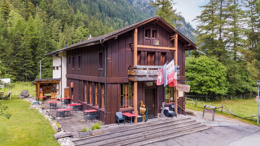 Chalet style building with outdoor seating surrounded by dense green forest and mountains.
