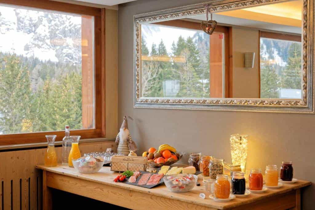 Breakfast buffet with fruit, jams, and cold cuts, overlooking snowy alpine forest through window.