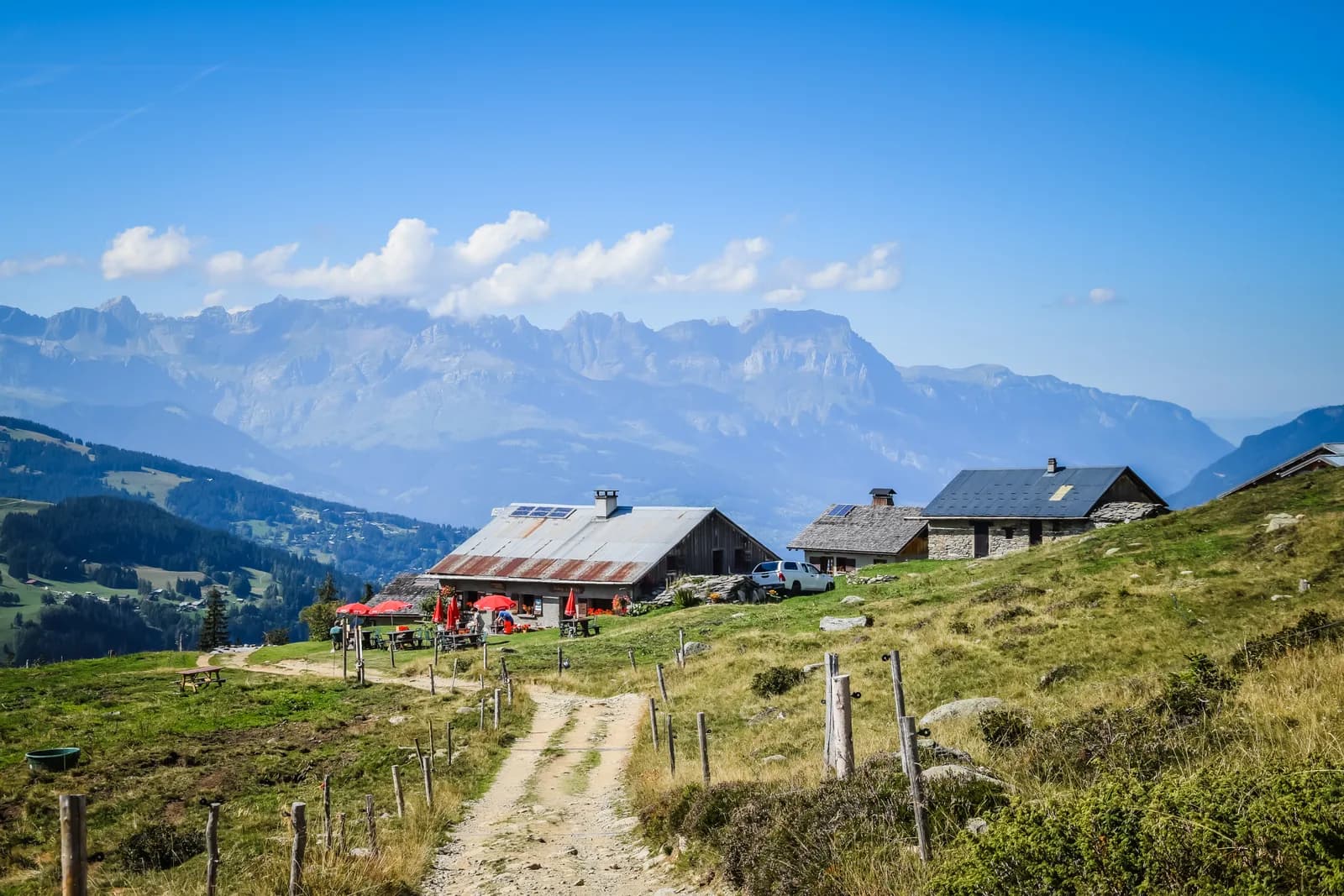 Mountain refuge with outdoor seating on grassy slope overlooking blue mountains under clear sky