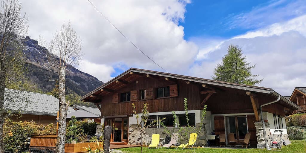 Chalet in Gite-les-Melezes with wooden facade and mountain backdrop under cloudy sky.
