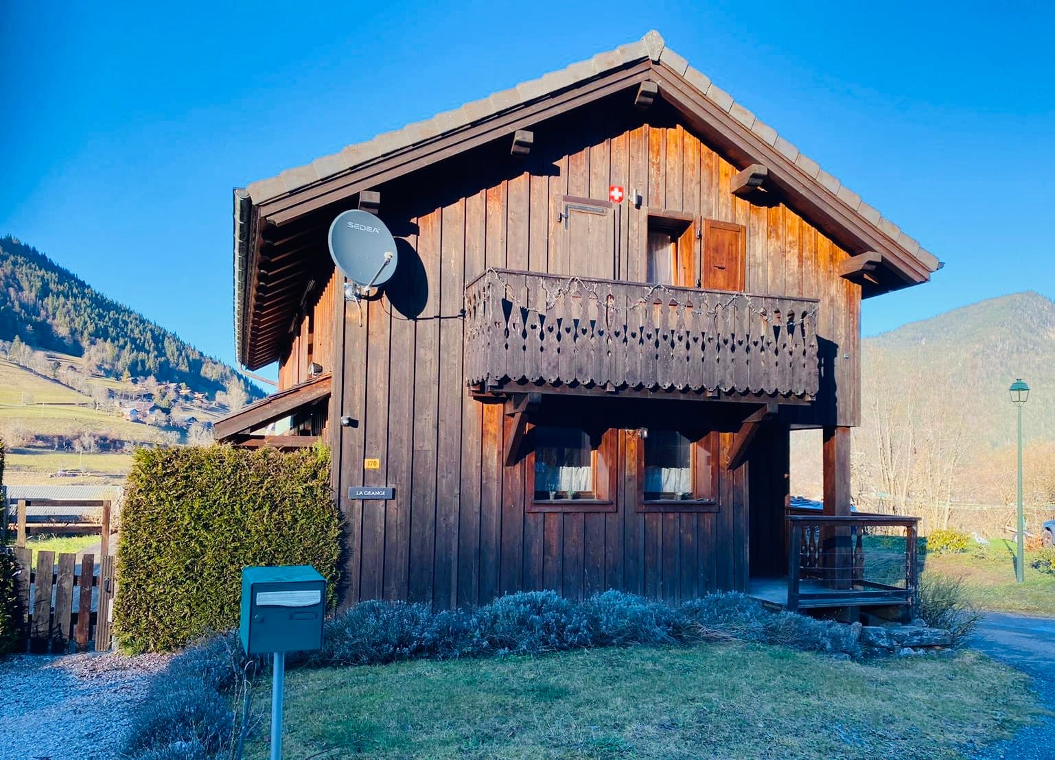 Wooden chalet named La Grange with balcony, set against a backdrop of forested mountains.