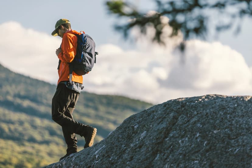 African American man hiking down a mountain with a backpack in an orange coat with ski area trails off in the distance in Vermont, New England, USA.
