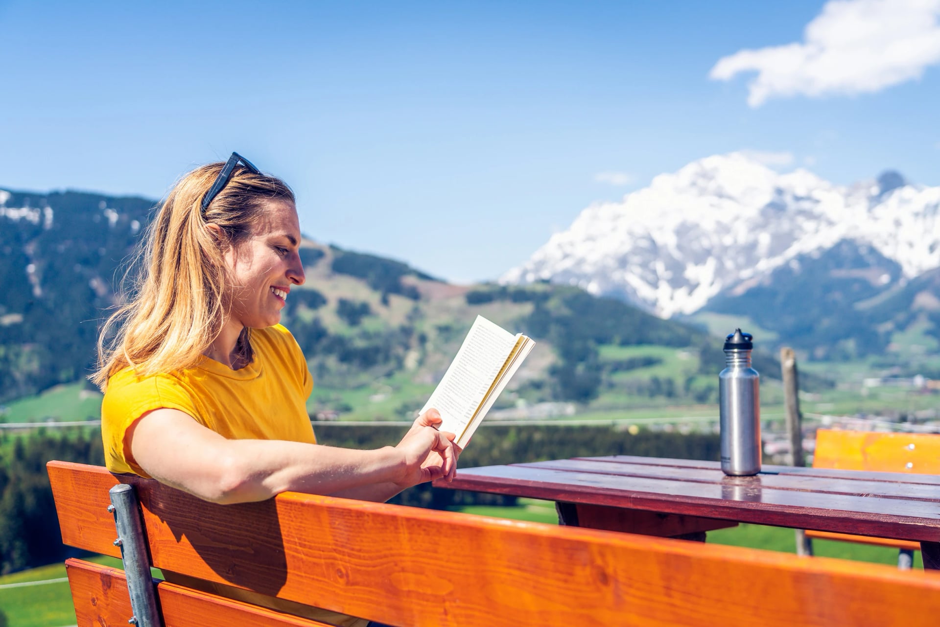 Young beautiful and happy girl reading a book on a bench, mountain range and greenery in the background.