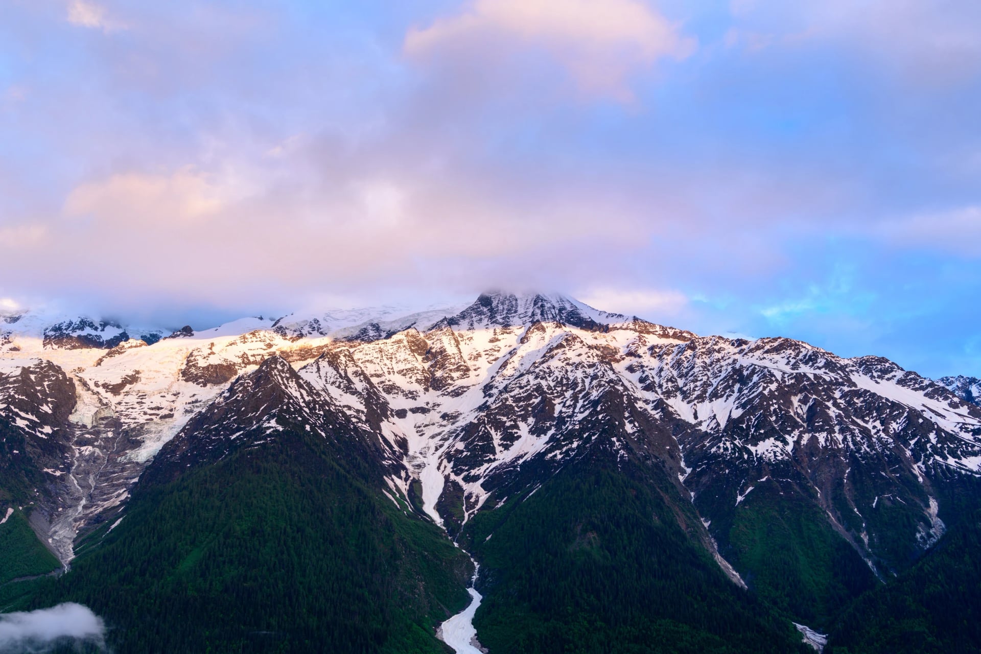 Warm morning light illuminates the rugged, snow-streaked peaks of the Mont Blanc massif, with patches of green forest below and pastel clouds overhead.