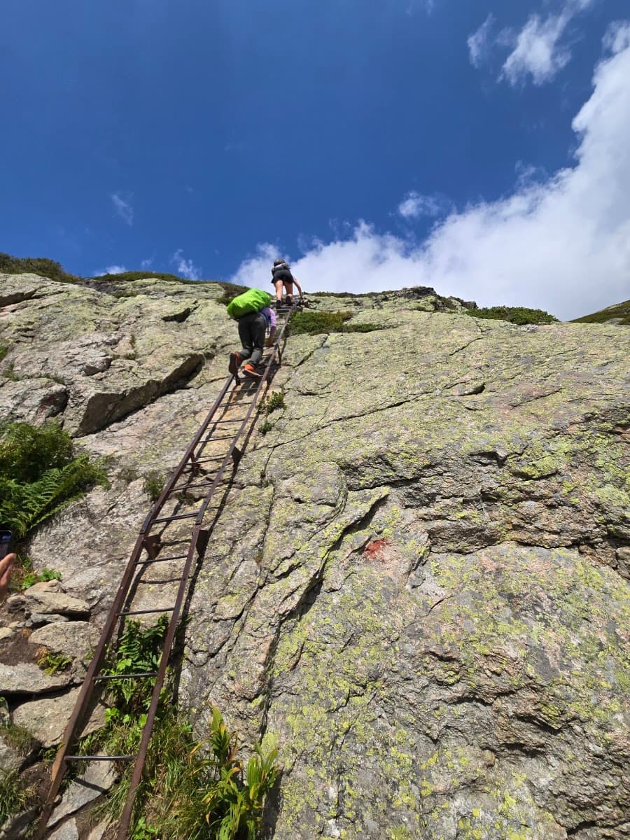 Hikers climbing a steep metal ladder on a rocky mountain slope under a blue sky